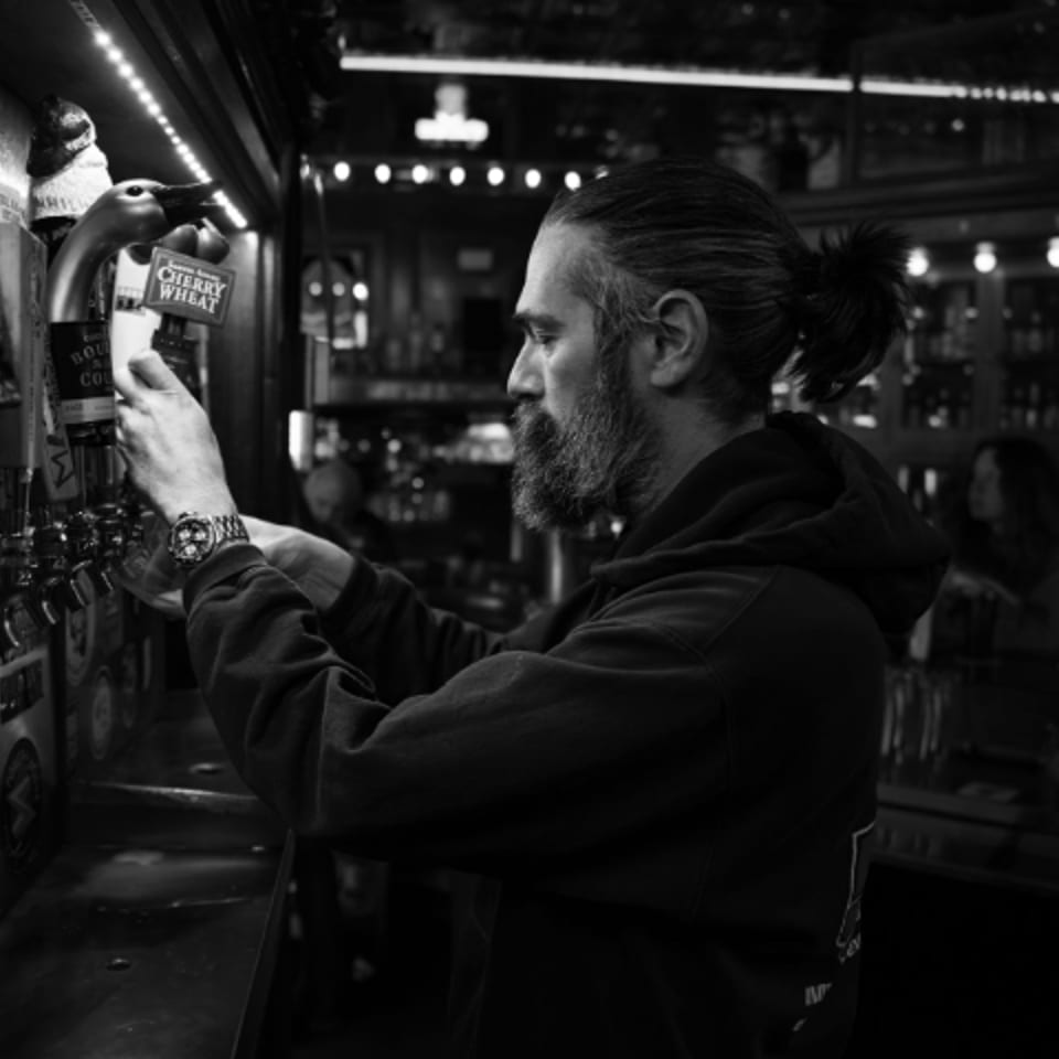 Bearded bartender with a ponytail pours beer from a tap in a dimly lit bar.