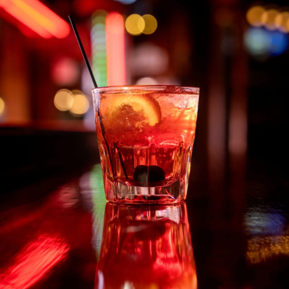 Red cocktail with citrus garnish and cherry in a rocks glass, reflecting on a bar counter amid colorful blurred lights.