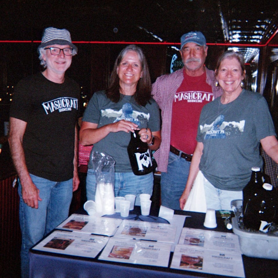 Four people in Mashcraft Brewing shirts stand behind a table with beer bottles and flyers.