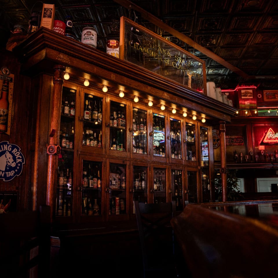 A dimly lit bar interior with a large wooden liquor cabinet, neon signs, and a dark bar top.