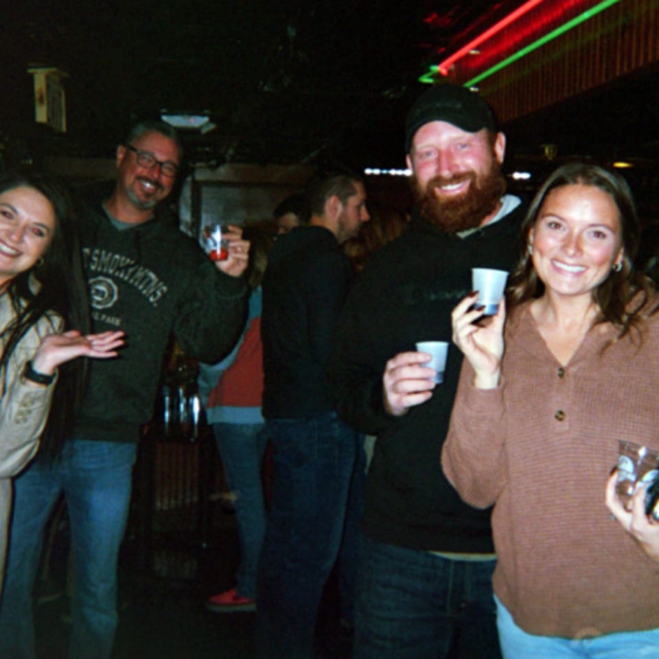 Friends smile and hold drinks at a lively, dimly lit bar with colorful neon lighting.