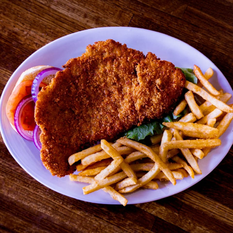 Crispy breaded fish fillet served with French fries, lettuce, tomato, and red onion on a white plate.