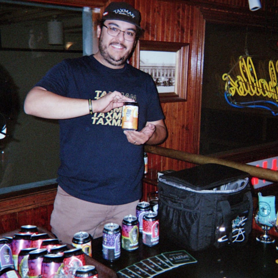 Smiling man in a Taxman shirt holds a beverage can behind a bar surrounded by colorful cans and a cooler.
