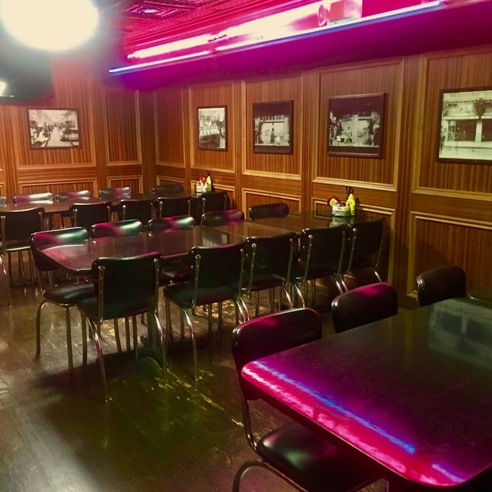A dimly lit dining room with wood-paneled walls, black tables, and chairs, illuminated by neon lights.