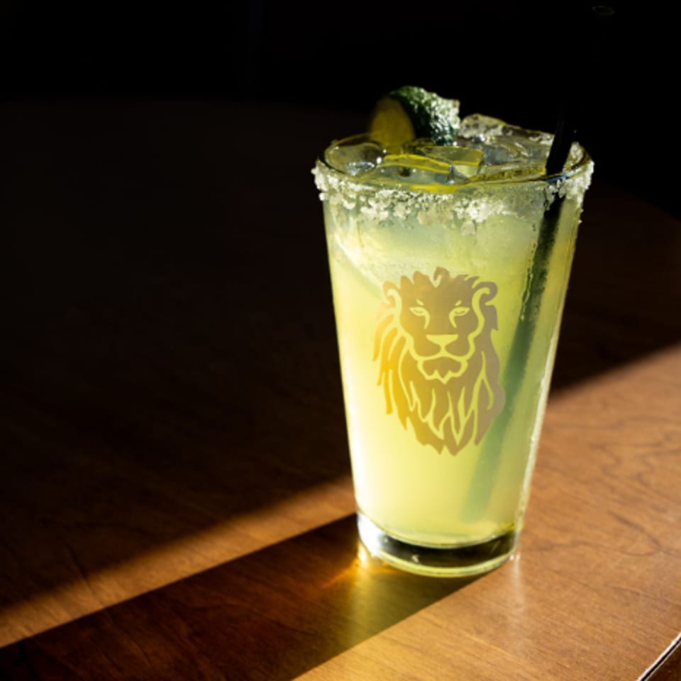 Yellow citrus cocktail with a salt-rimmed glass, lime garnish, straw, and lion logo on a sunlit wooden table.