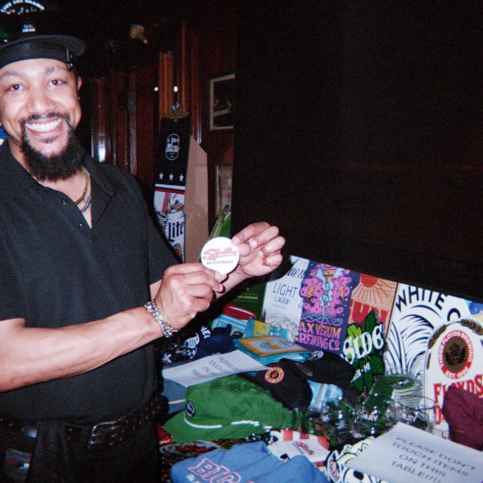 Smiling man in a black hat holds a restaurant button beside a colorful merchandise display in a dim bar.