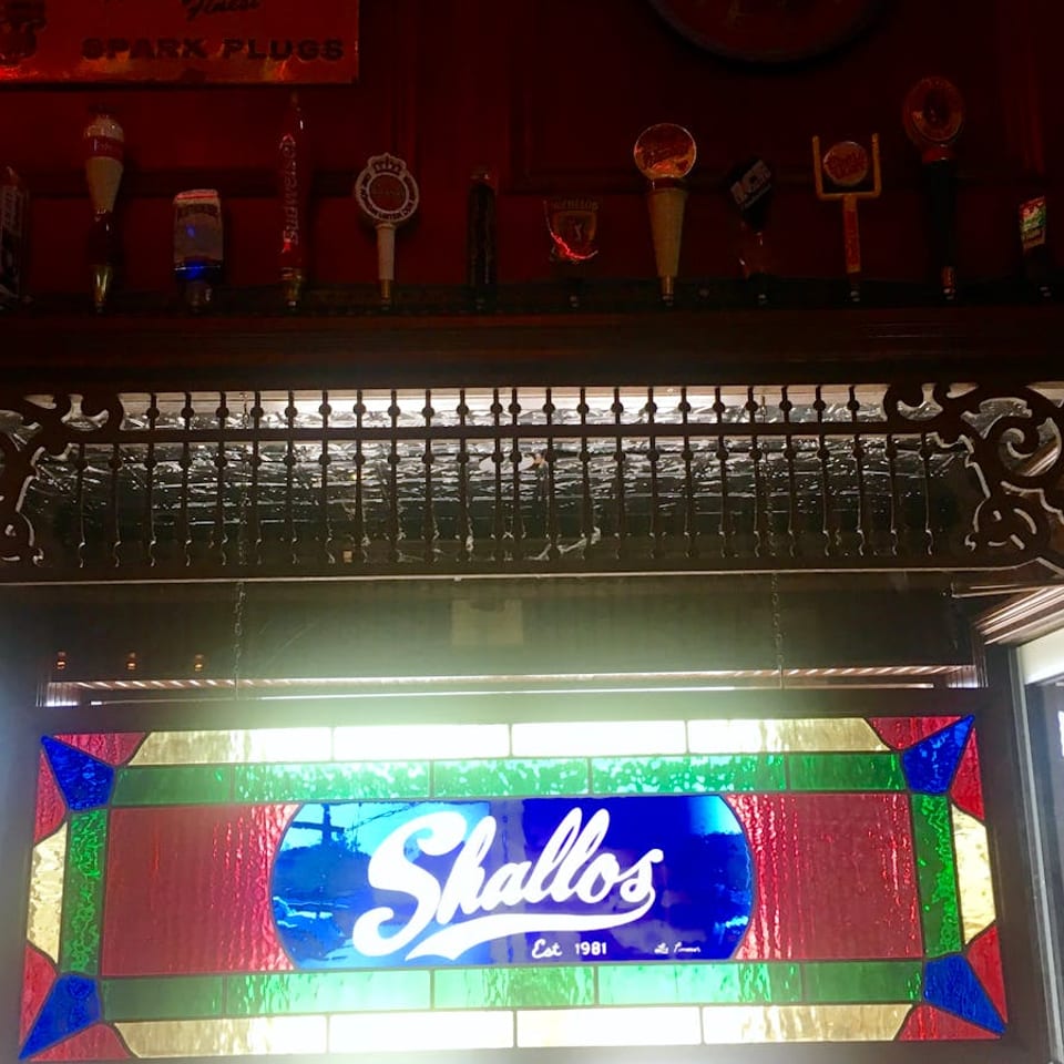 A stained glass sign for "Shallos Est. 1981" hangs below a shelf displaying various beer tap handles.
