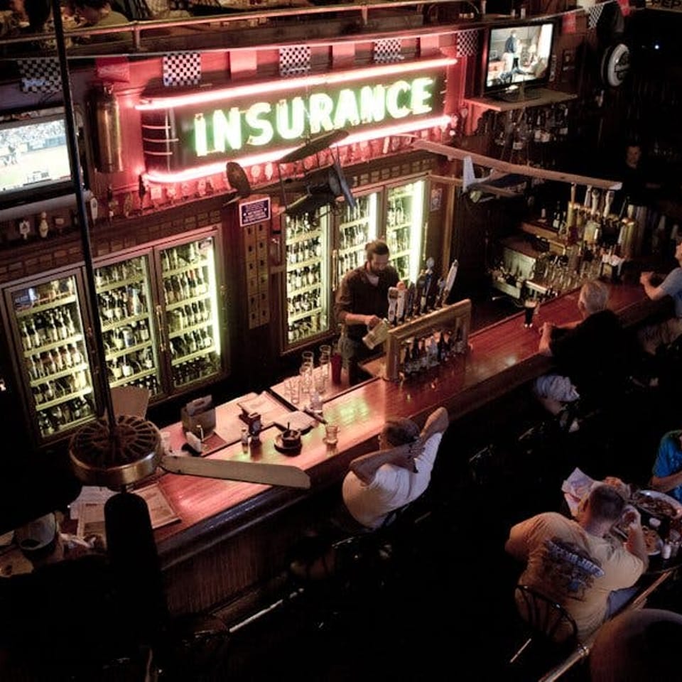 Overhead view of a dimly lit bar with a neon "INSURANCE" sign, bartender, and patrons.