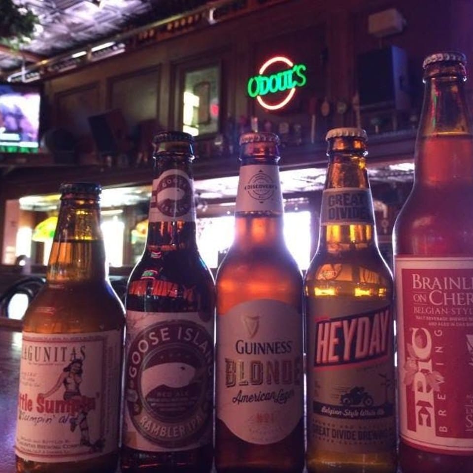 Five diverse beer bottles, including Guinness Blonde and Goose Island, lined up in a dimly lit bar.