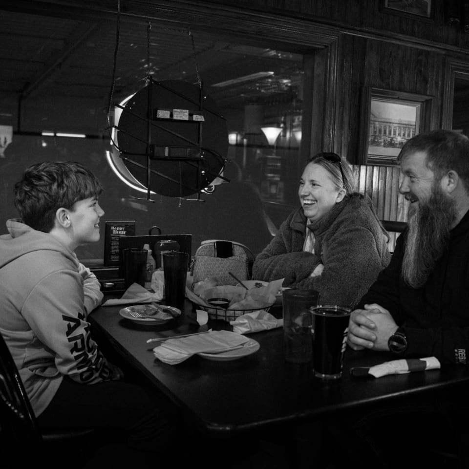 Three people laughing and talking at a dark restaurant table with drinks and food.