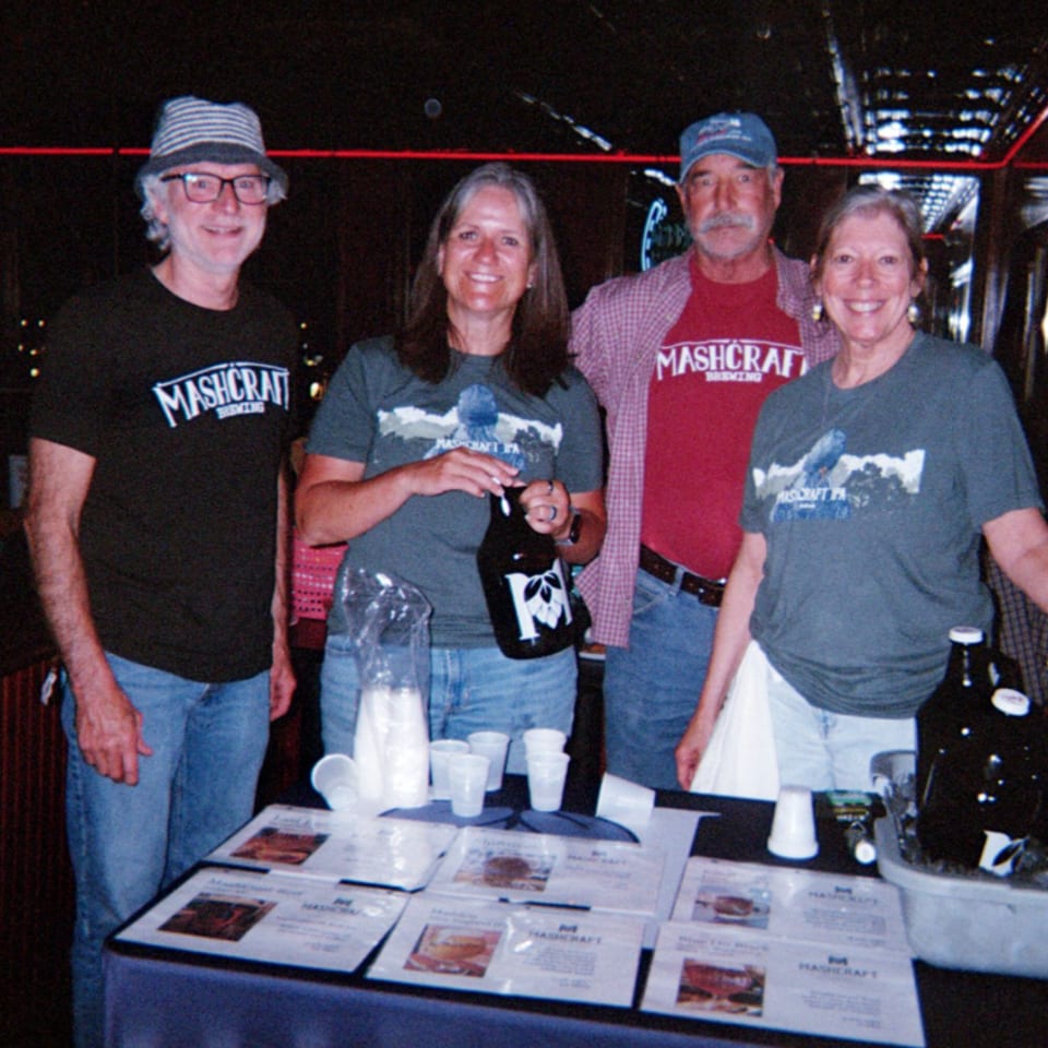 Four MashCraft Brewing representatives pose behind a table displaying beer bottles, plastic cups, and promotional materials in a dim venue.
