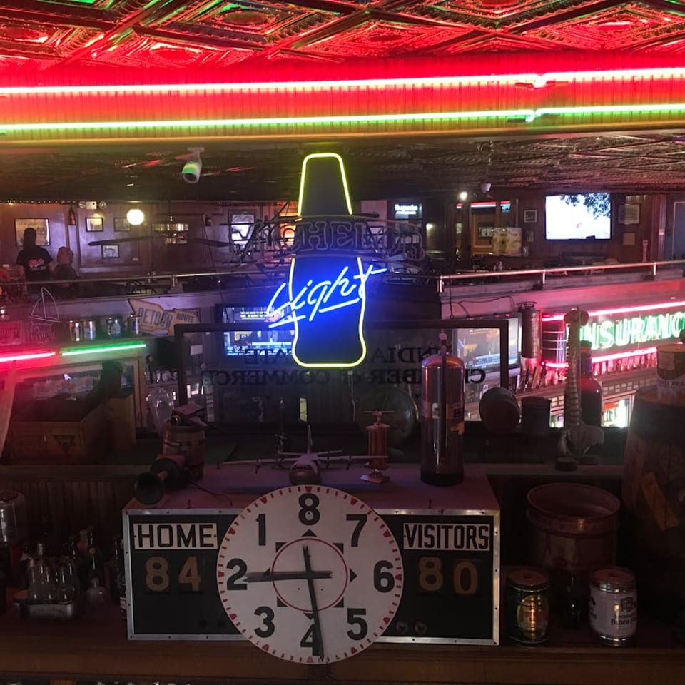 Neon lights illuminate a lively bar interior with a vintage scoreboard and various decorations.