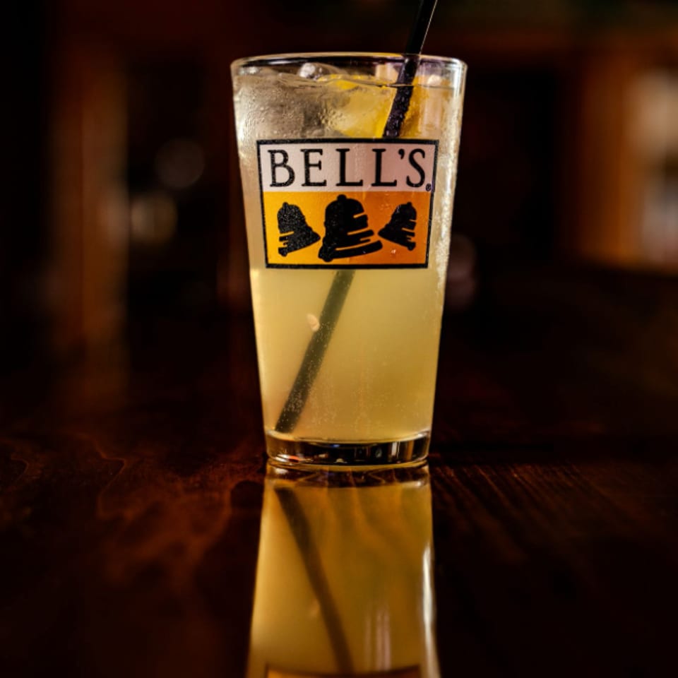Pale golden cocktail with ice, lemon garnish, and straw in a Bell’s-branded glass on a reflective wooden bar.