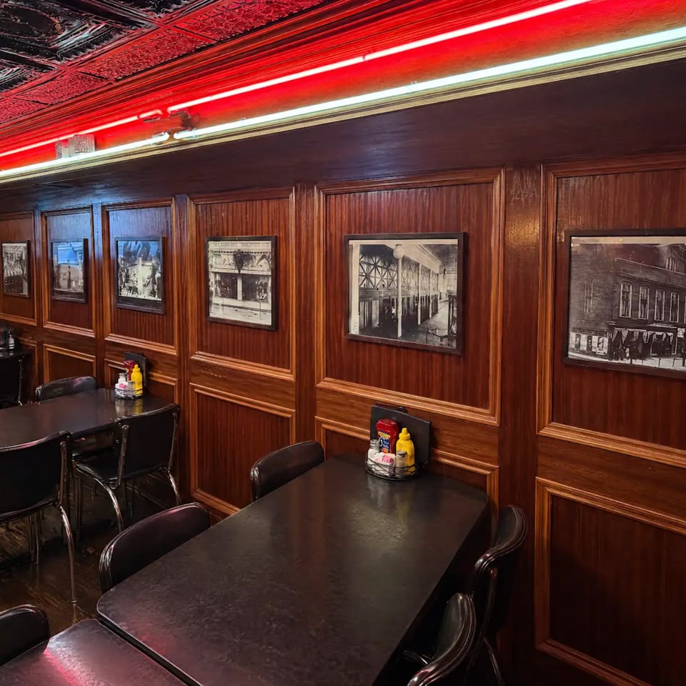 Restaurant dining room with dark wood paneling, black tables and chairs, framed historic photographs, and red neon lighting.