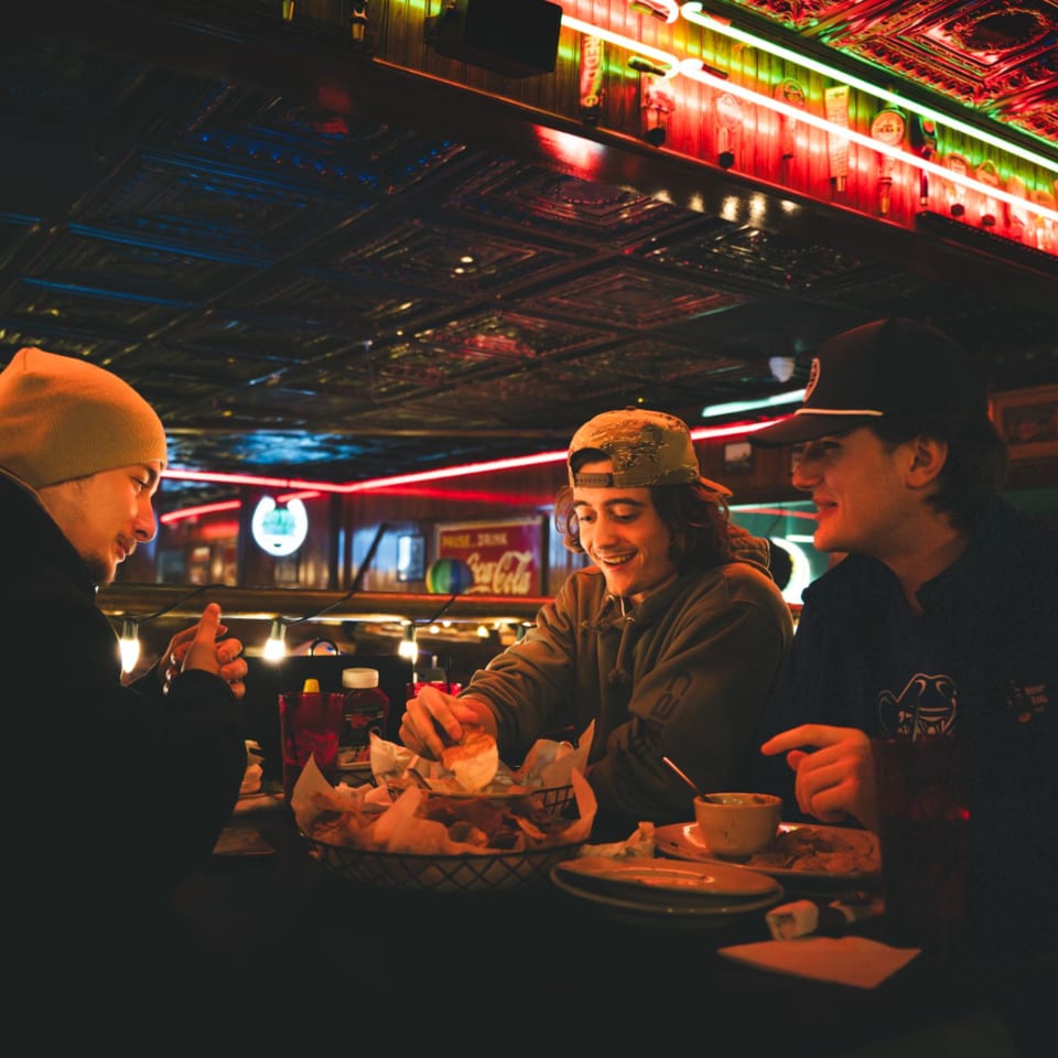 Three young men enjoying food and drinks at a dimly lit bar with neon signs.