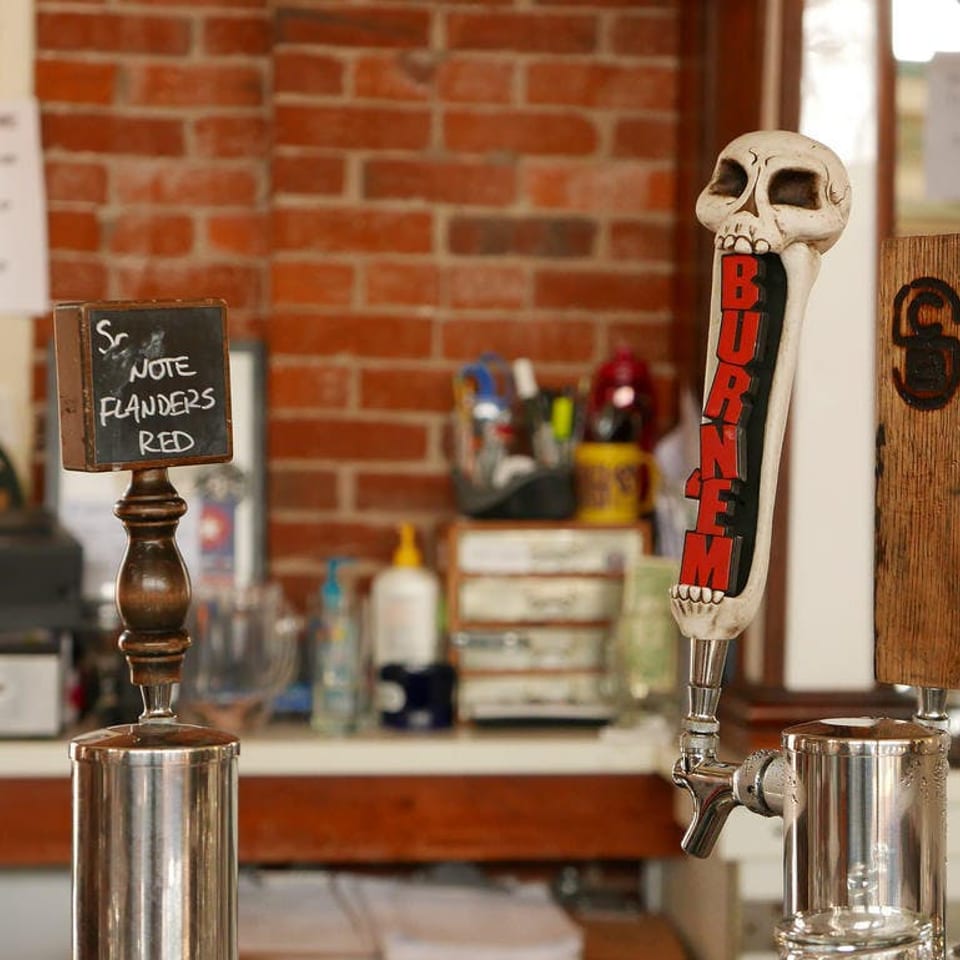 Three beer taps on a bar, one with a skull handle, against a brick wall.