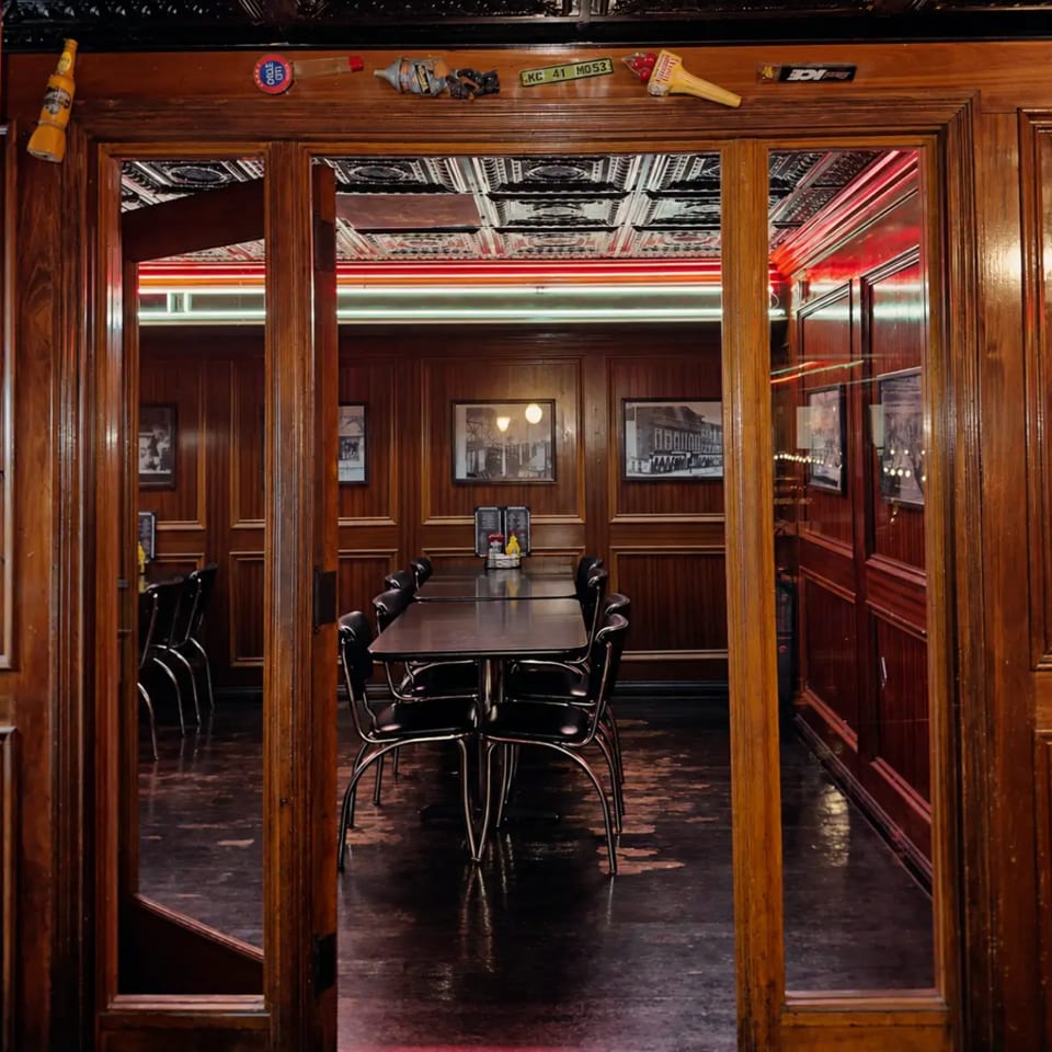 Vintage restaurant dining room with wood paneling, mirrored doors, neon lighting, framed artwork, and black tables and chairs.