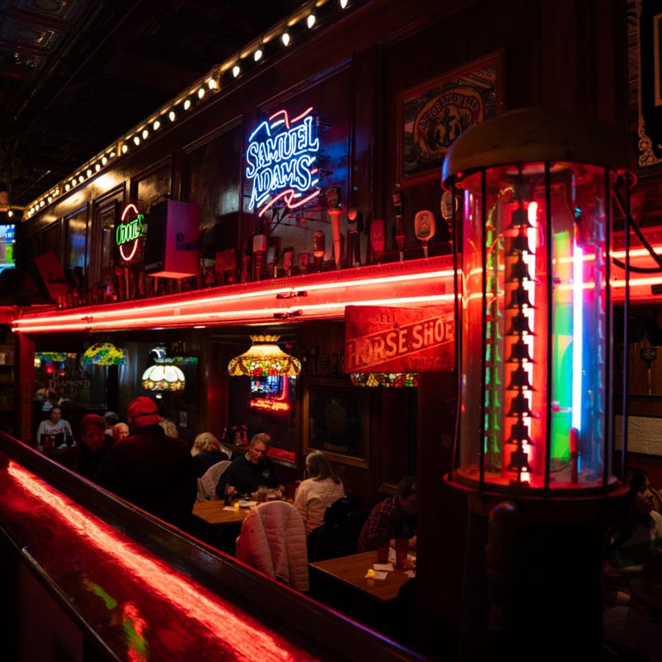 Dark, lively bar interior with red neon lights, patrons seated at tables, and a glowing vintage gas pump.