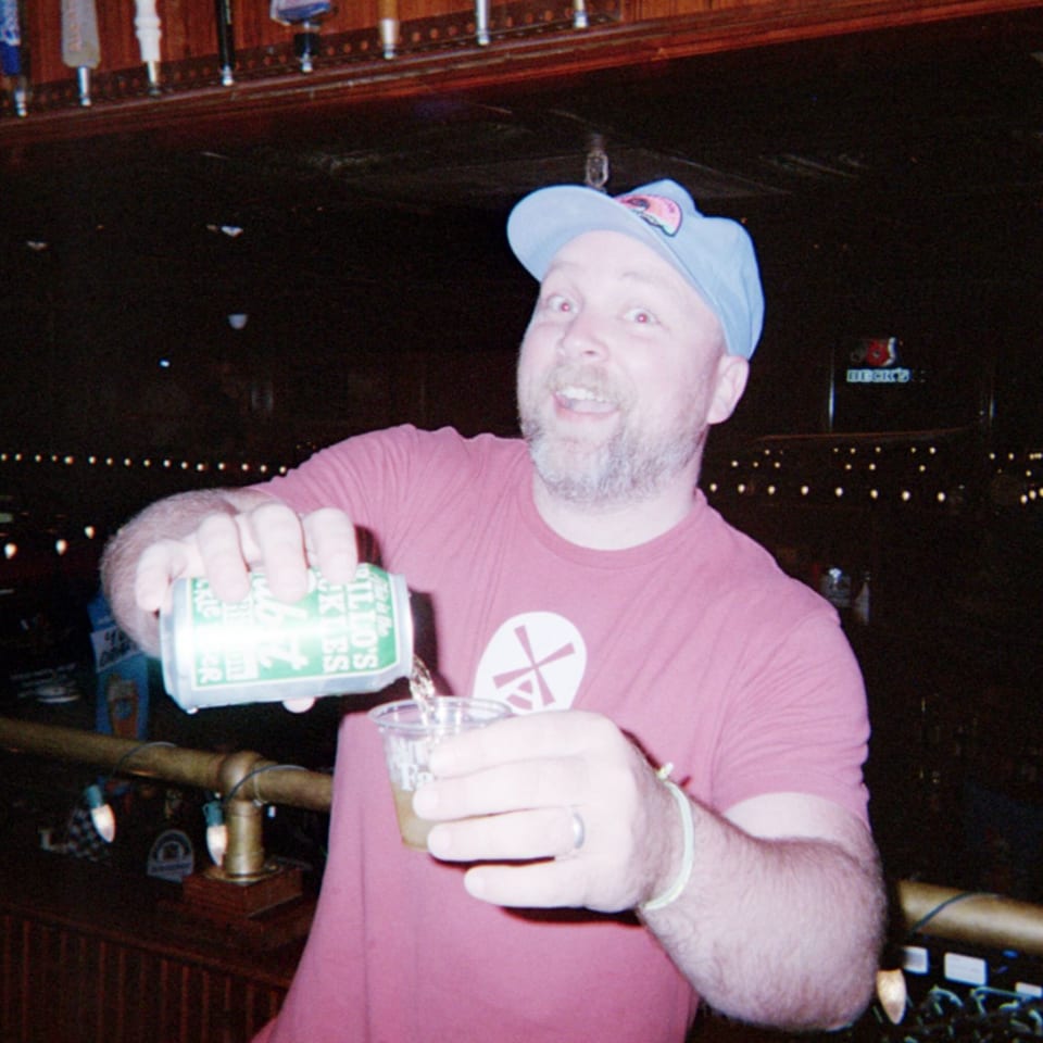 Bartender pours soda from a can into a glass at a dimly lit bar with string lights.