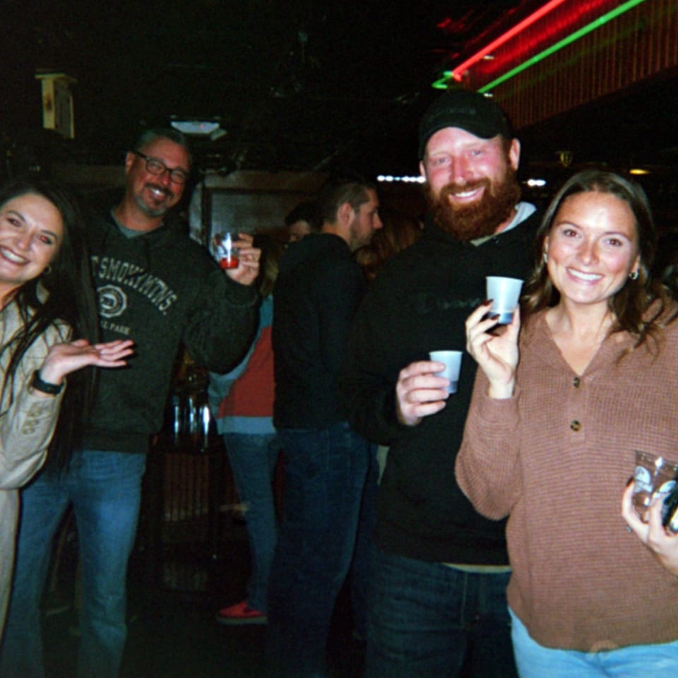 Four smiling people holding drinks, enjoying a lively evening in a dimly lit bar with neon lights.