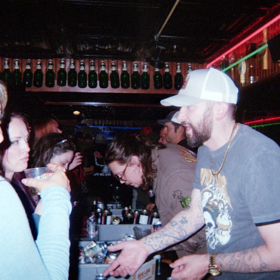 Woman holding a drink beside a bearded man in a cap at a crowded, neon-lit bar.