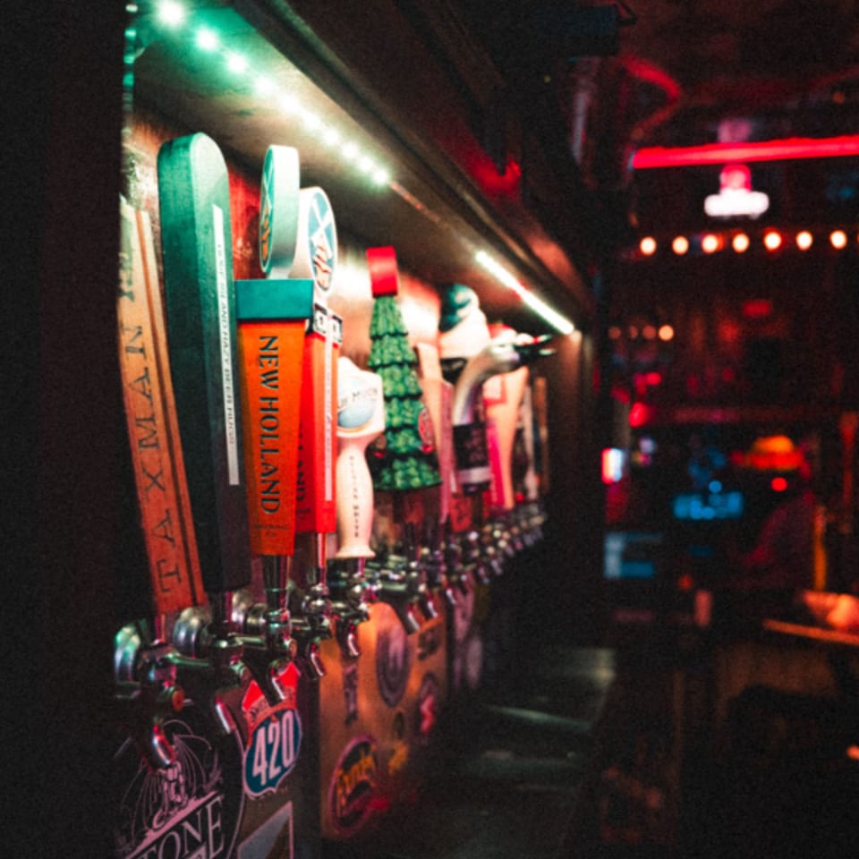 Row of colorful craft beer taps glowing under green and red lights along a dimly lit bar.