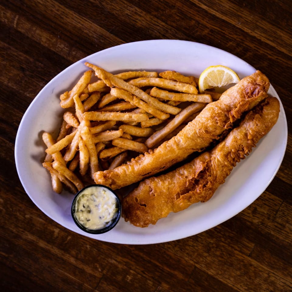 Plate of golden fish and chips with two battered fish fillets, fries, lemon wedge, and tartar sauce on a wooden table.